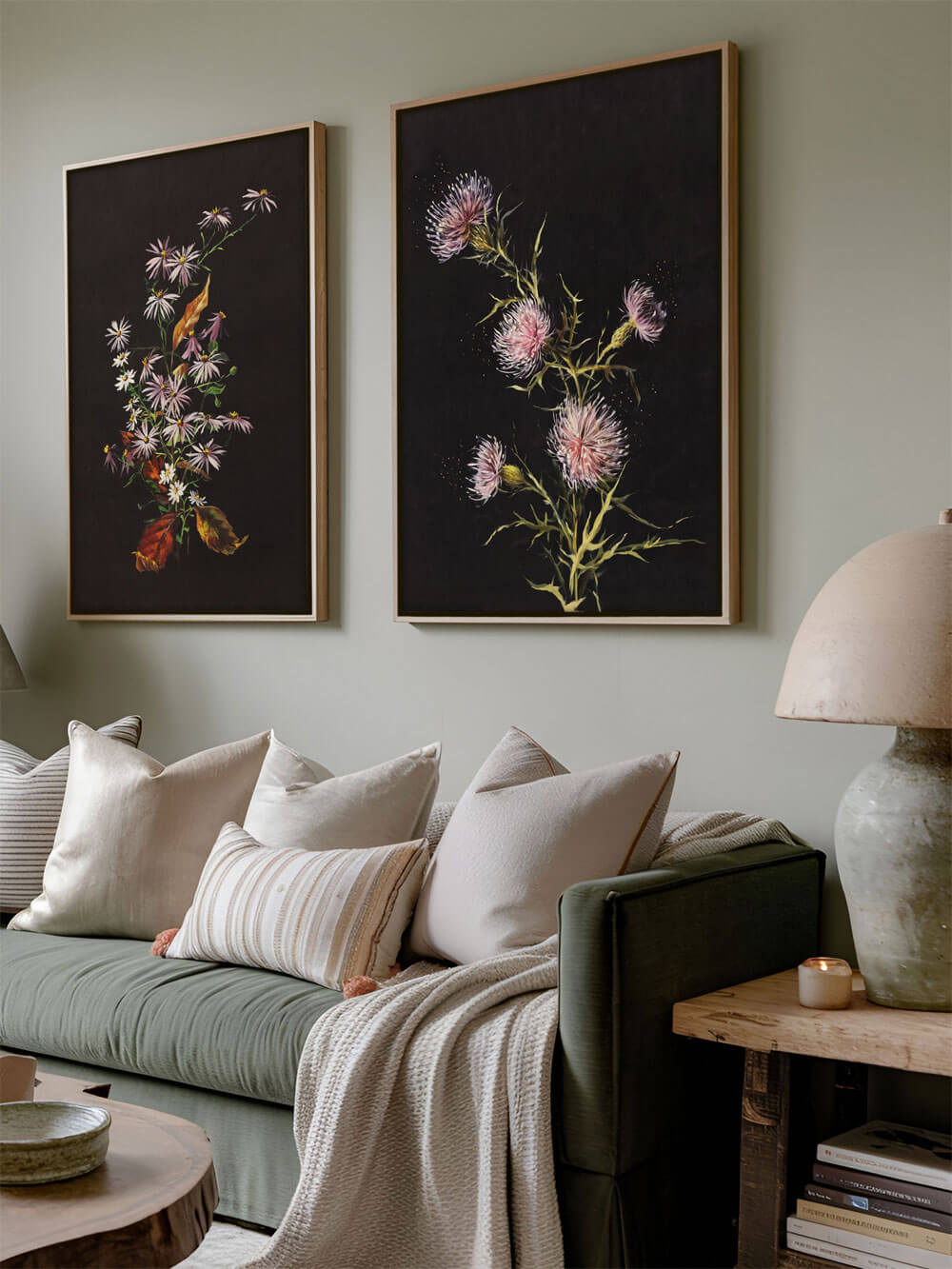 Zwei große Kunstdrucke mit floralen Motiven hängen nebeneinander über einem olivgrünen Sofa. Die linke Darstellung zeigt feine, pastellfarbene Blumen mit Blättern und dezenten gelben Akzenten, während das rechte Bild stachelige rosa Blüten darstellt. Der Hintergrund beider Bilder ist tiefdunkel, wodurch die farbigen Details der Pflanzen strahlen. Die beige Kissen und die Lampe ergänzen den natürlichen, ruhigen Stil des Raumes.