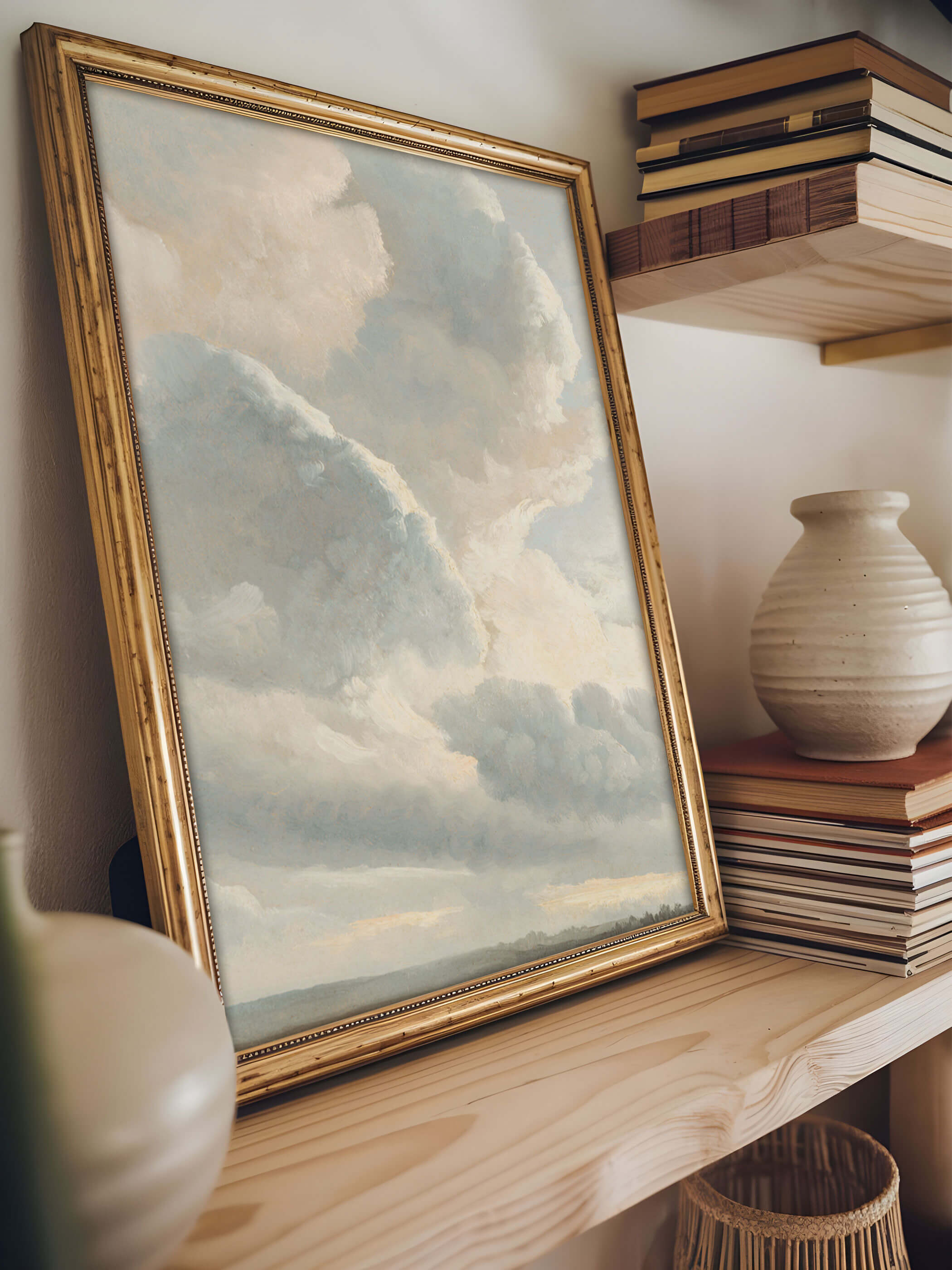 Vintage Wolken-Kunstdruck mit goldener Rahmung, aufgestellt auf einem Holzregal neben Büchern und einer Vase. Zarte Wolken in sanften Farben schaffen eine ruhige Dekoration.